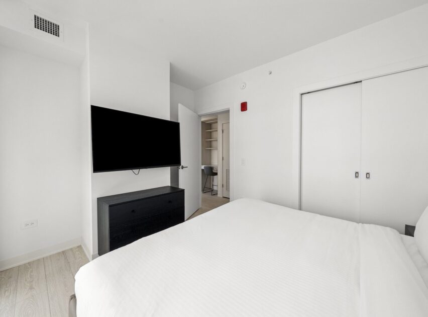 Minimalist bedroom with a neatly made white bed, wall-mounted TV above a black dresser, open door revealing a small office nook, and white sliding closet doors. The room features light wood floors and white walls.