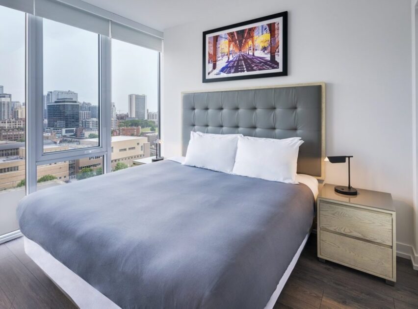 A modern bedroom with a large bed featuring a gray headboard and sheets, two white pillows, a wooden nightstand with a lamp, large windows with city views, and a framed cityscape photo on the wall above the bed.