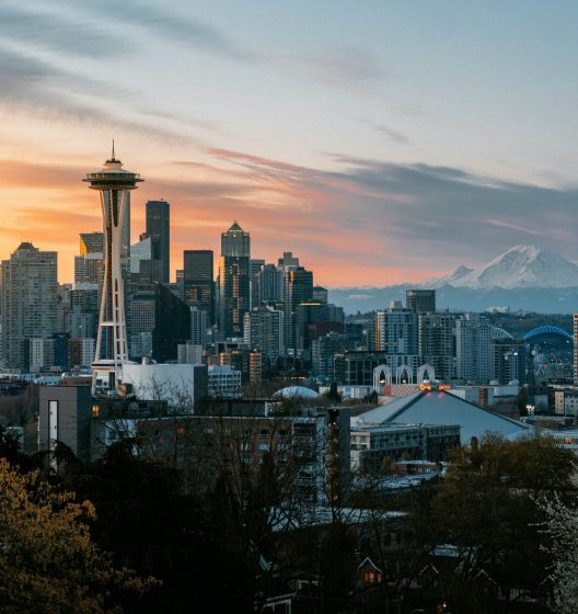 Seattle skyline at sunset with the Space Needle in the foreground, downtown skyscrapers, and Mount Rainier visible in the background under a partly cloudy sky.