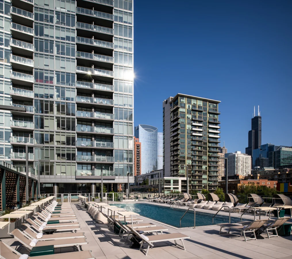 A modern rooftop pool with rows of lounge chairs is surrounded by high-rise apartment buildings against a clear blue sky, with a city skyline visible in the background.