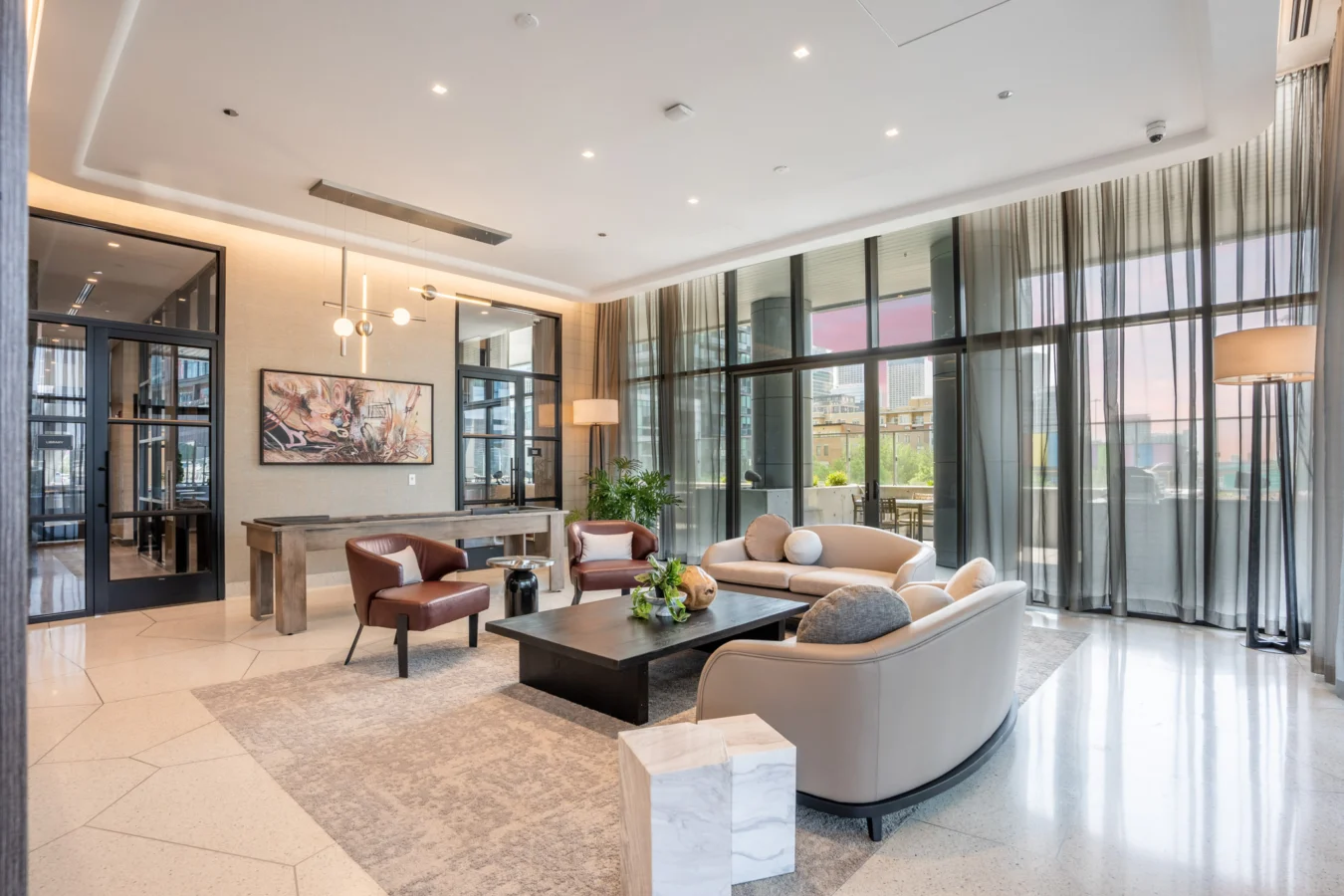 Modern living room with large windows, sheer curtains, neutral-toned sofas, armchairs, a black coffee table, floor lamps, artwork, indoor plants, and a view of an outdoor patio at sunset.