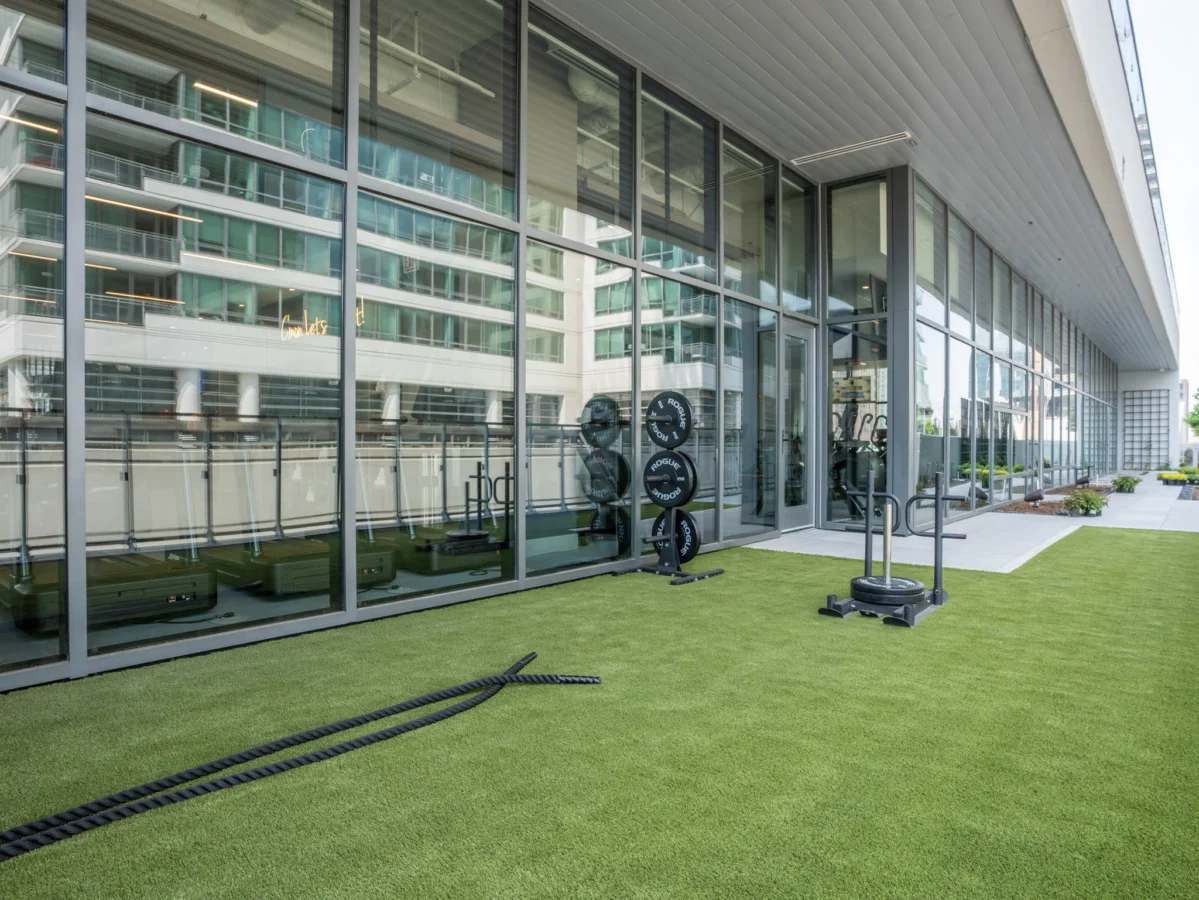Outdoor gym area with artificial grass, battle ropes, weights, and fitness equipment next to a modern building with large glass windows reflecting surrounding structures.