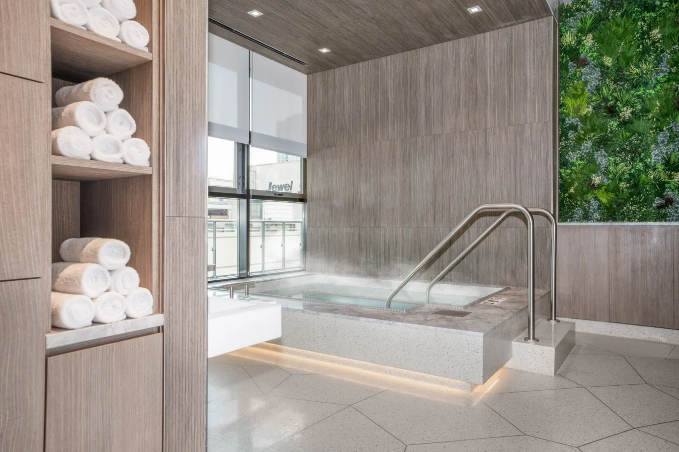 A modern spa room with a steaming indoor hot tub, wooden walls, shelves stacked with rolled white towels, and a vertical green plant wall for decoration. Natural light enters through large windows.