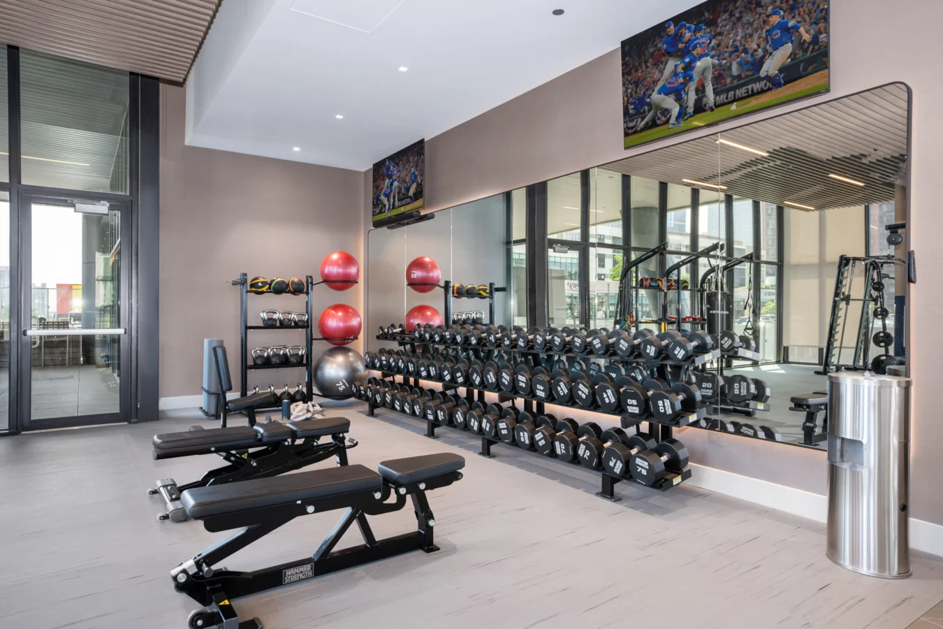 Modern gym with benches, dumbbells, exercise balls, medicine balls, and a large mirror. There are two TVs on the wall showing a football game. Floor-to-ceiling windows provide natural light.