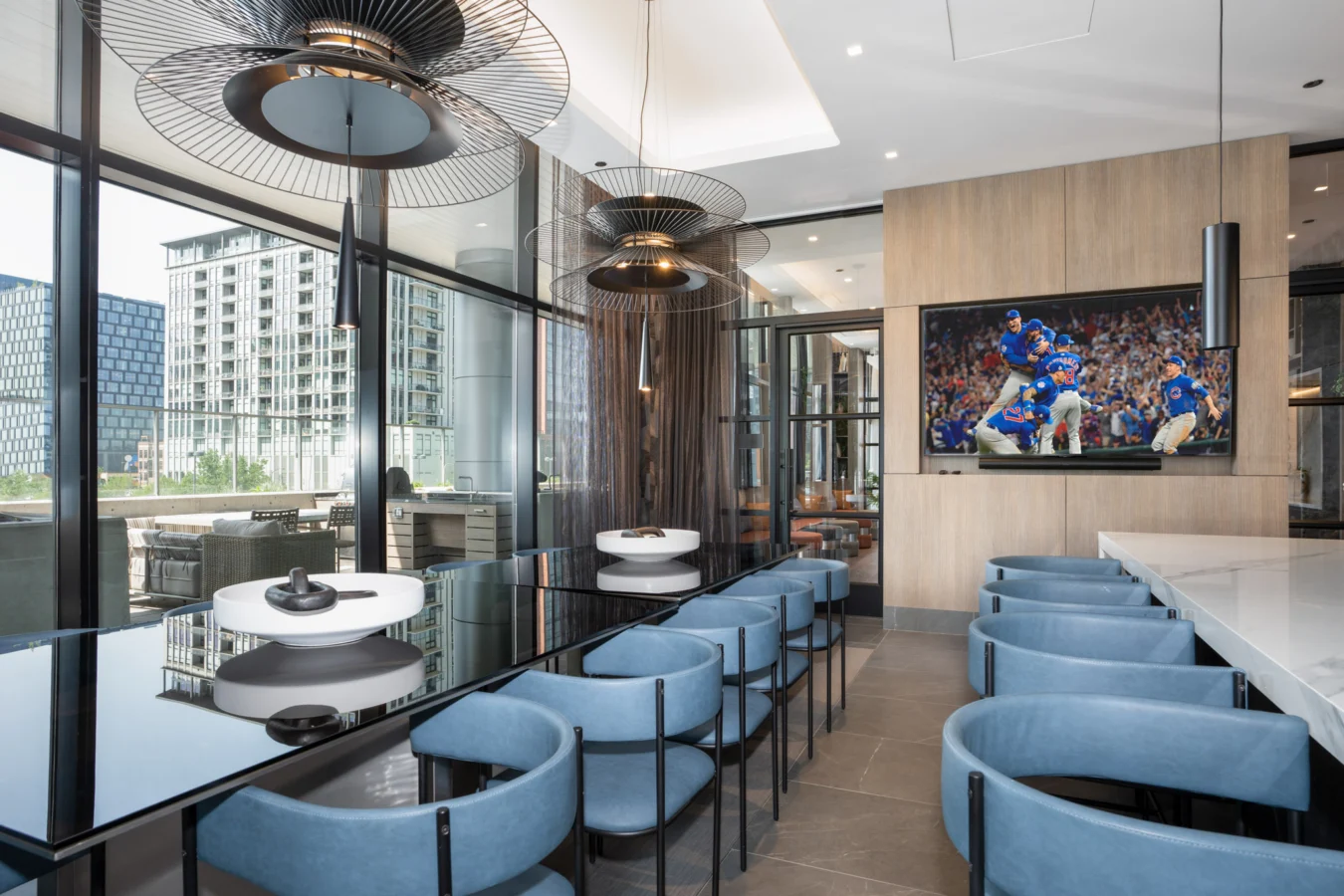 Modern lounge with blue chairs, high-gloss black tables, and large pendant lights. A TV on the wood-paneled wall displays a football game. Floor-to-ceiling windows reveal city buildings and an outdoor patio.