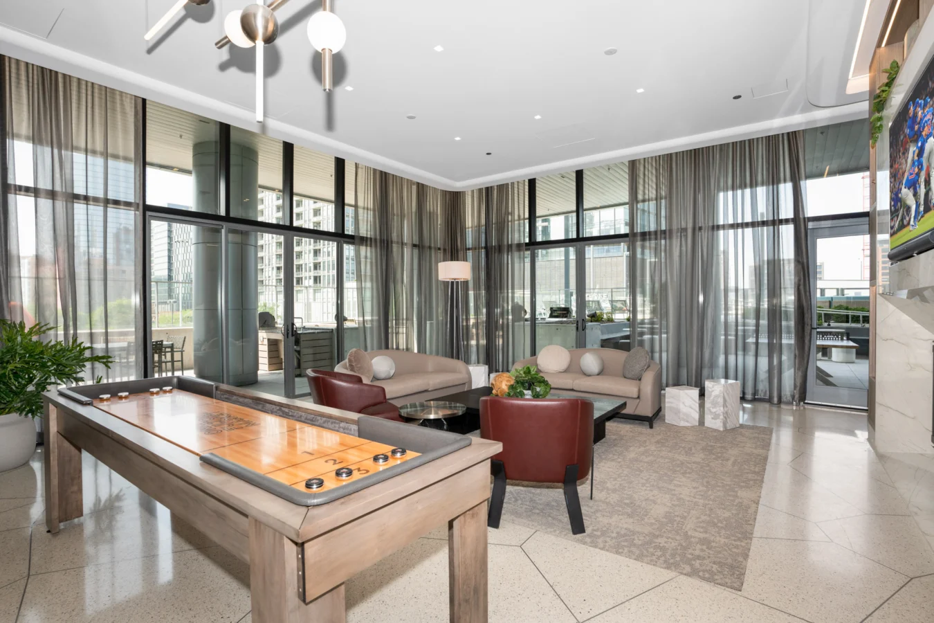 Modern lounge area with floor-to-ceiling windows, sheer curtains, two sofas, two red chairs, a coffee table with plants, a shuffleboard table, and a view of a patio and city buildings outside.
