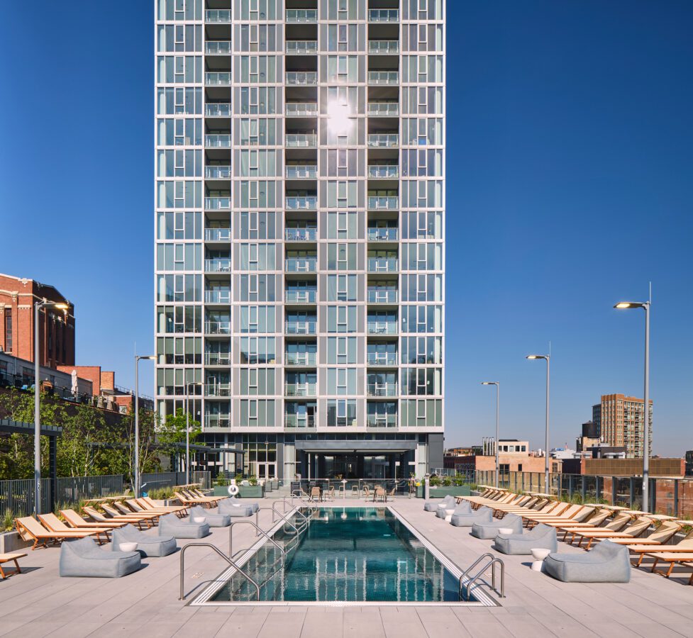 A modern high-rise apartment building with a central outdoor pool surrounded by lounge chairs and umbrellas on a sunny day, set against a clear blue sky and urban skyline.