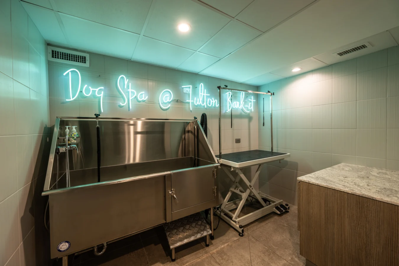 A modern dog spa with a stainless steel washing station, grooming table, and a neon sign reading Dog Spa @ Fulton Bank. The room has tiled walls, a countertop, and soft overhead lighting.