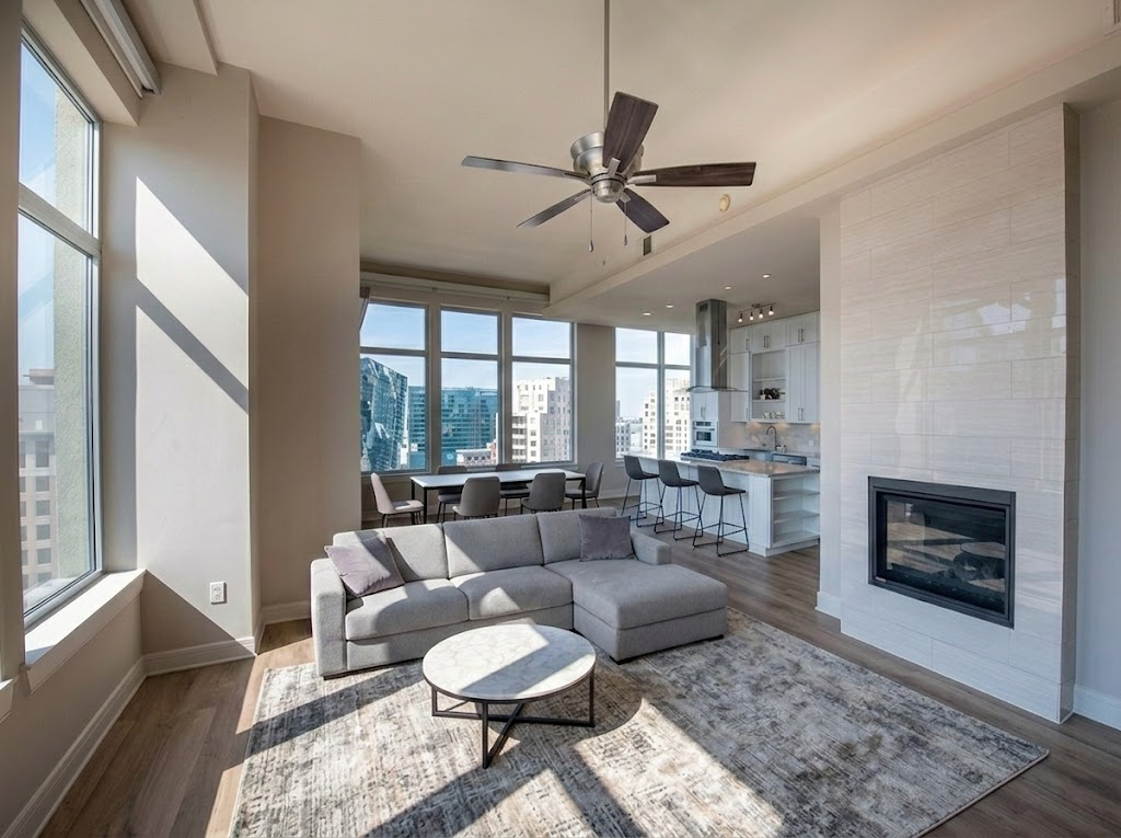 Modern, sunlit living room with large windows, a gray sectional sofa, round coffee table, area rug, ceiling fan, fireplace, and an open kitchen with barstools and city views in the background.