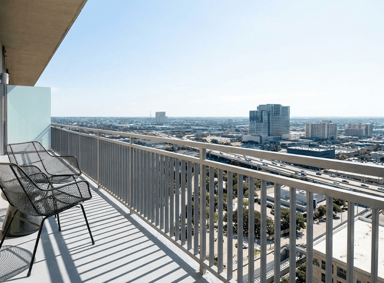A high-rise balcony with two black metal chairs overlooks a city skyline, featuring tall buildings, roads, and a clear blue sky in the background.