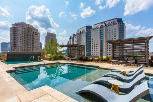 Rooftop pool area with lounge chairs in the water, cabanas, and city buildings in the background under a blue sky with clouds.