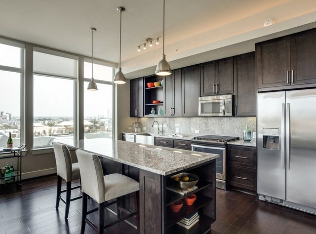 Modern kitchen with dark wood cabinets, stainless steel appliances, granite island with two upholstered chairs, open shelving with dishes, and large windows letting in natural light with a city view.
