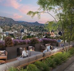 People relax on outdoor sofas on a rooftop terrace with city views, lush greenery, and purple flowers. The Hollywood sign and hills are visible in the background under a colorful evening sky.
