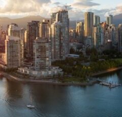 A view of downtown Vancouver with modern high-rise buildings, a waterfront park, and a small sailboat on calm water, with mountains in the background under a partly cloudy sky.