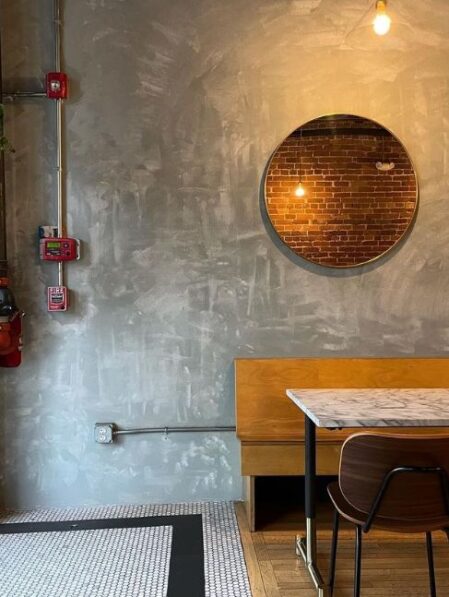 A modern cafe corner with a marble table, wooden chair, and bench against a textured gray wall. A round mirror reflects a brick wall and hanging light bulb. Fire safety equipment is mounted on the left.