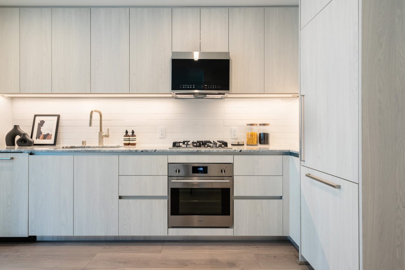 Modern kitchen with light wood cabinets, stainless steel oven, gas cooktop, built-in range hood, and minimalist decor including a framed photo, vases, and jars on a marble countertop against a white tile backsplash.