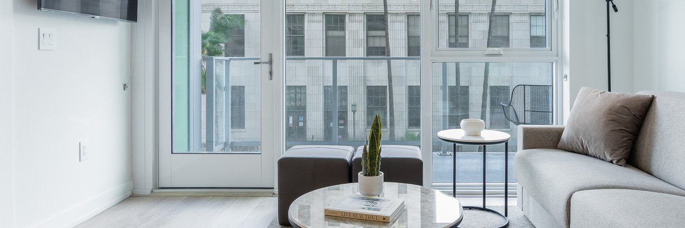 Modern living room with a light gray sofa, marble coffee table with a small plant and books, two ottomans, and floor-to-ceiling windows showing a city building outside. Minimalist decor in neutral tones.