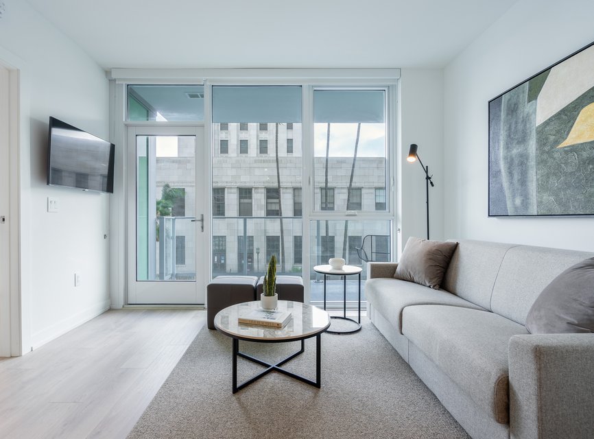 Modern living room with a gray sofa, round coffee tables, wall-mounted TV, large abstract art, floor lamp, and floor-to-ceiling windows revealing a city building outside.