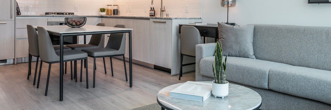 Modern apartment interior with a light gray sofa, round coffee table with a plant and books, dining table with four chairs, and a kitchen area with light wood cabinets and a bowl of fruit.