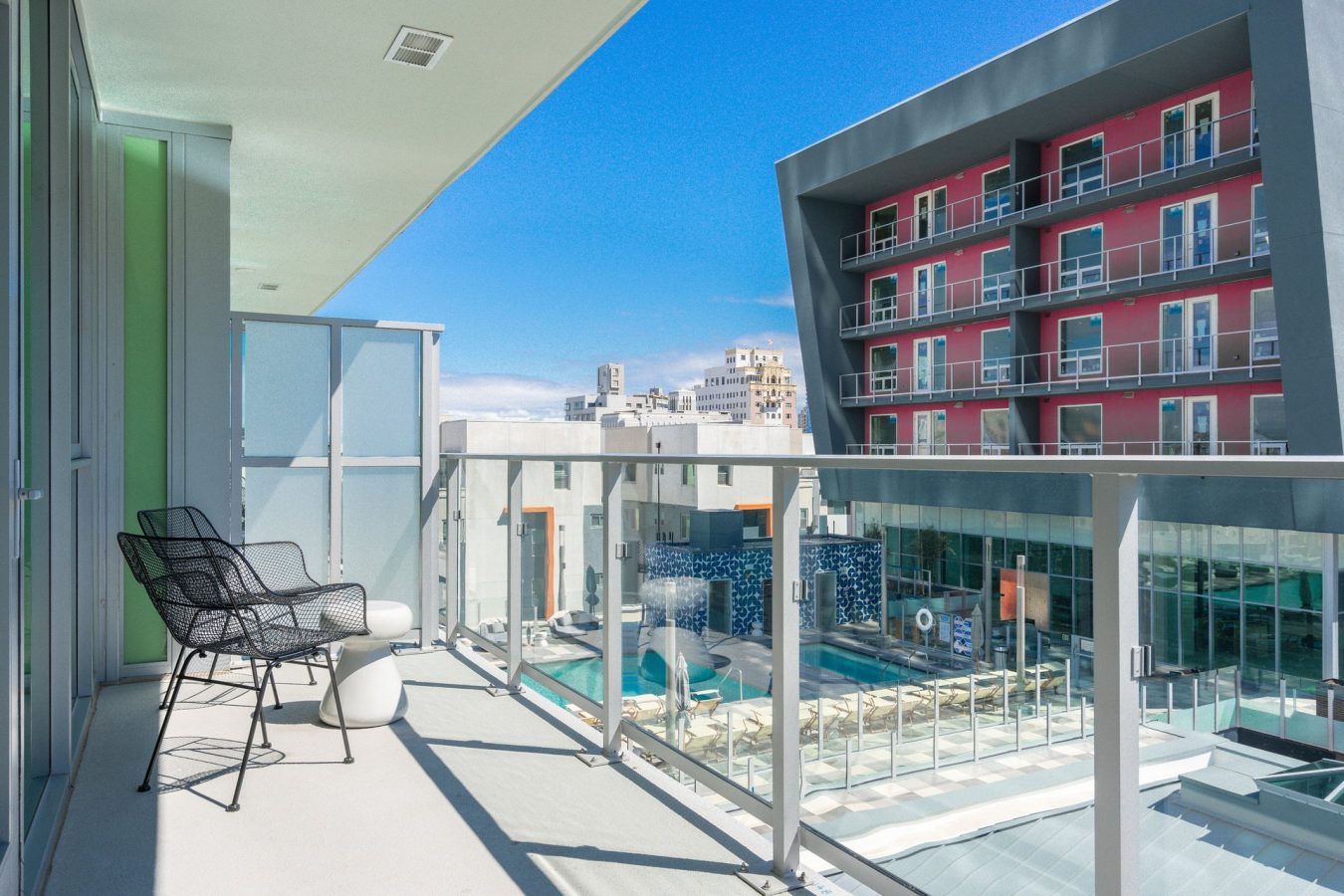 Sunny apartment balcony with glass railing, two black chairs, and a small white table overlooks a modern building with red accents and a swimming pool below. Blue sky is visible in the background.