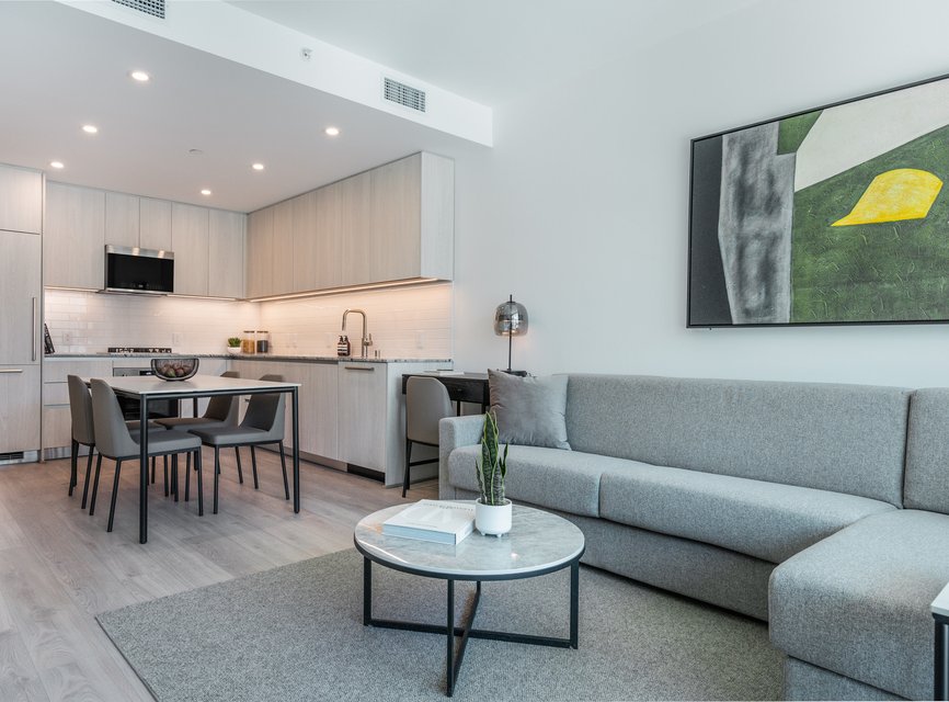 Modern open-plan living area with a gray sofa, round coffee table, small dining table with four chairs, and a minimalist kitchen. Abstract art hangs on the white wall. The space is bright with neutral tones.