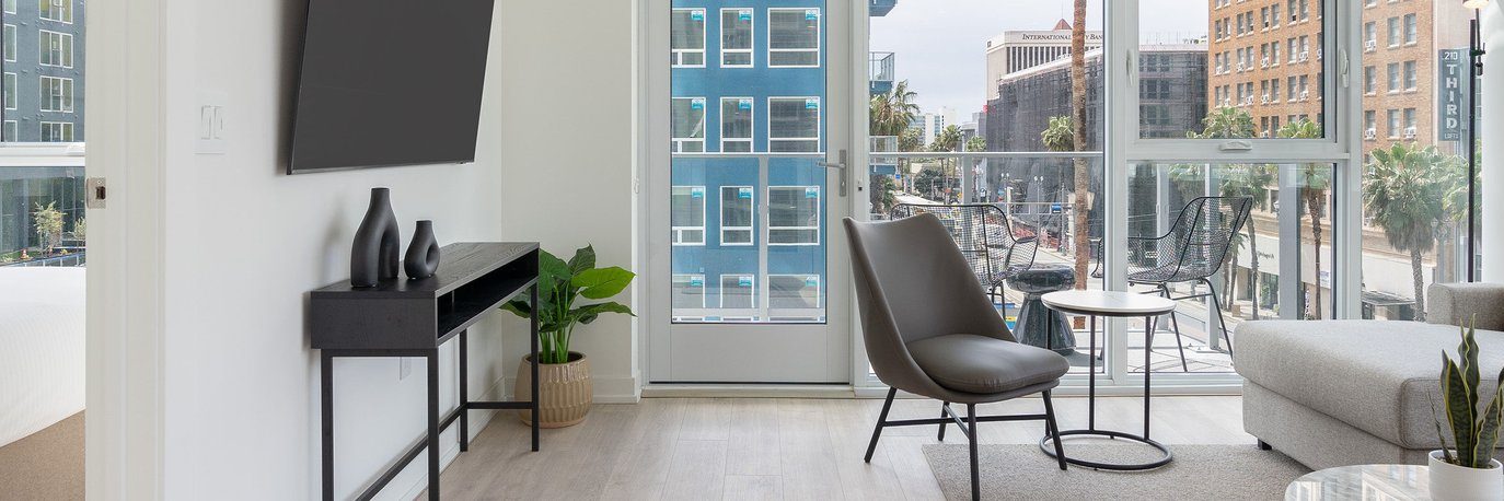 Modern living room with a gray chair, small round table, light sofa, black console table with vases, potted plant, and large windows showing a city street and tall buildings outside.