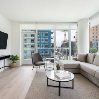 A modern, bright living room with large windows, a gray sectional sofa, a round coffee table, a black accent chair, and a potted plant. The space opens onto a balcony overlooking city buildings.
