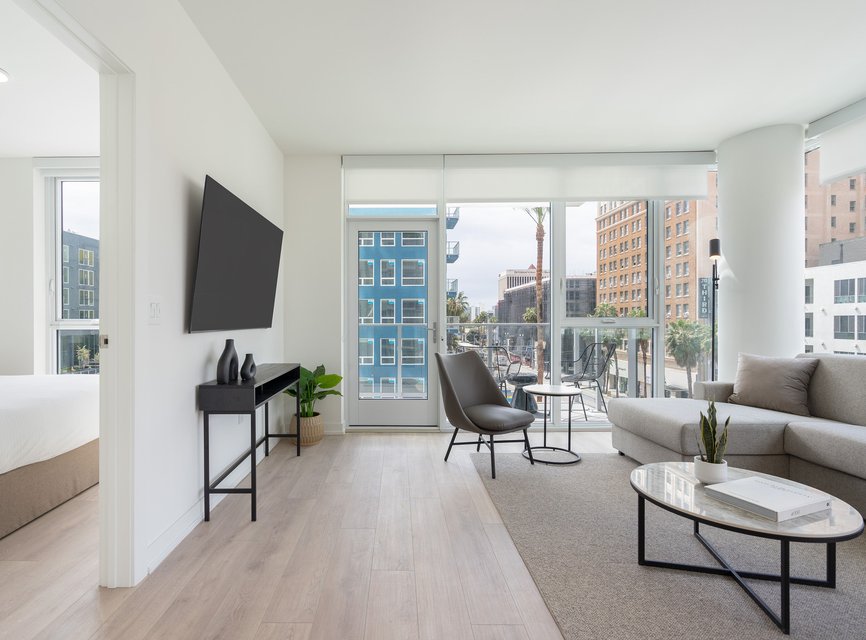 Modern, bright living room with large windows, light wood flooring, and neutral furniture. A mounted TV, gray sofa, chair, coffee table, and decor accents complete the minimalist, airy space. Balcony offers city views.
