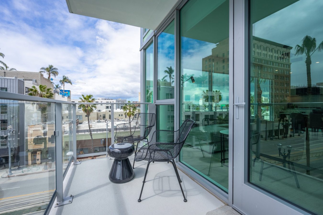 A modern balcony with glass railings, two black mesh chairs, and a small black table overlooks a city street lined with palm trees and contemporary buildings under a partly cloudy sky.