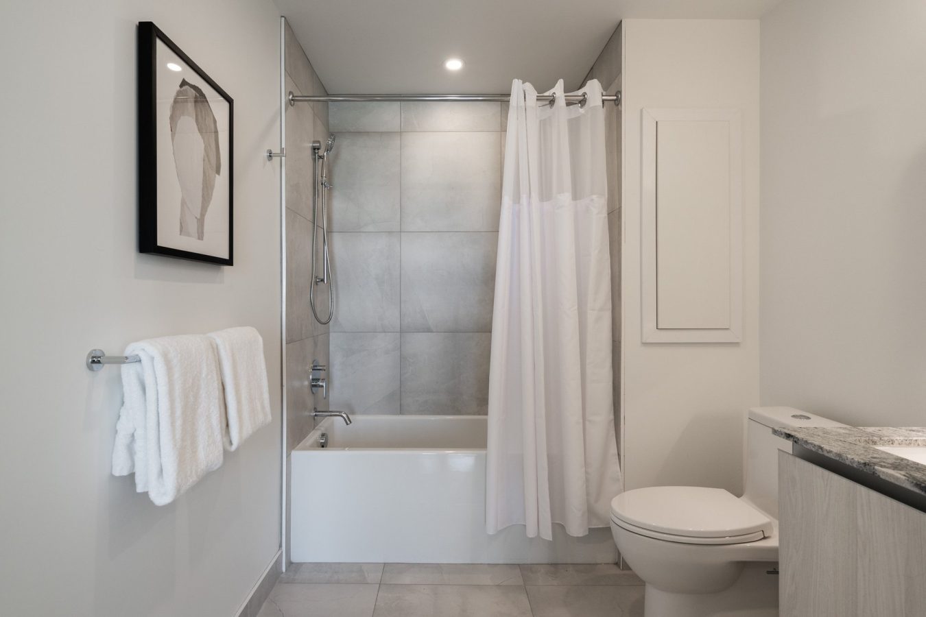 Modern bathroom with a white bathtub and shower, gray tile walls, white shower curtain, toilet, towel rack with white towels, wall art, and marble countertop. The room has a clean, minimalist design.