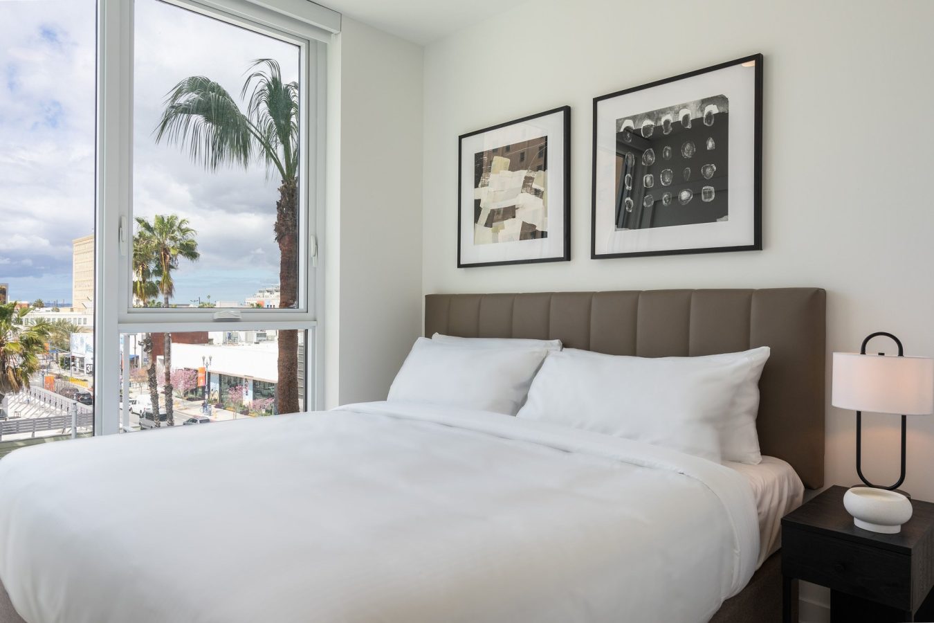 A modern bedroom with a neatly made bed, two abstract art pieces on the wall, a nightstand with a lamp, and a large window showing palm trees and buildings outside.