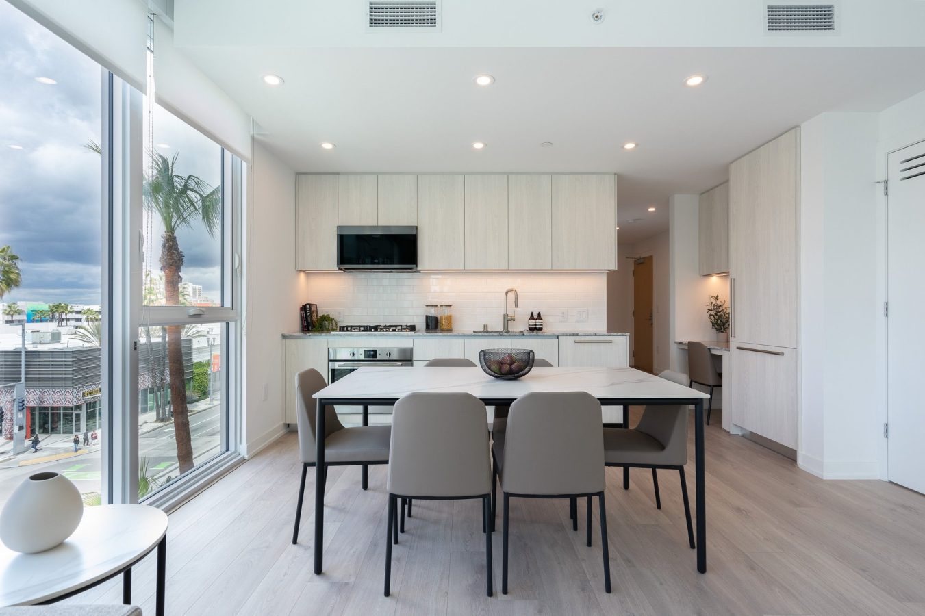 Modern kitchen and dining area with light wood cabinets, a white table set for four, large windows letting in natural light, and cityscape views with palm trees outside. Contemporary decor and minimalist design.