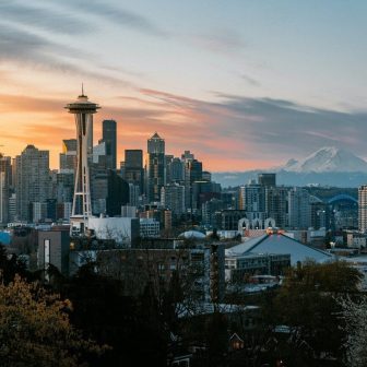 Seattle skyline at sunset featuring the Space Needle, various skyscrapers, and Mount Rainier in the background under a partly cloudy sky. Trees and the city’s sports arena are visible in the foreground.
