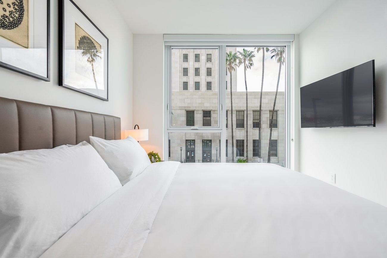 A bright, modern bedroom with a large bed, white bedding, brown upholstered headboard, wall art, a mounted TV, and a window with a city building view and palm trees outside.
