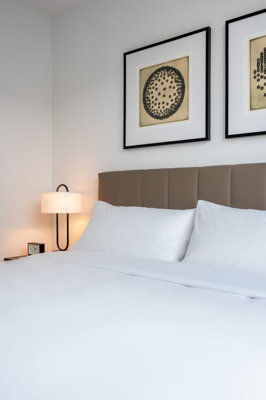 A neatly made bed with white linens and a beige headboard sits beside a nightstand with a lamp and clock. Two framed abstract art pieces hang on the light-colored wall above the bed.