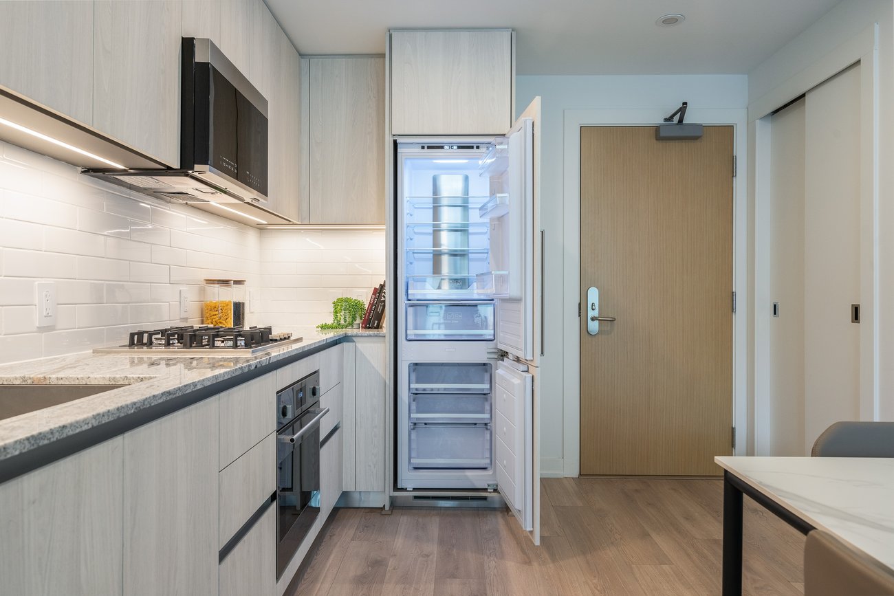 Modern kitchen with light wood cabinets, a granite countertop, built-in stainless steel appliances, and an open refrigerator. The space features a light wood floor, white tile backsplash, and a wooden entry door in the background.