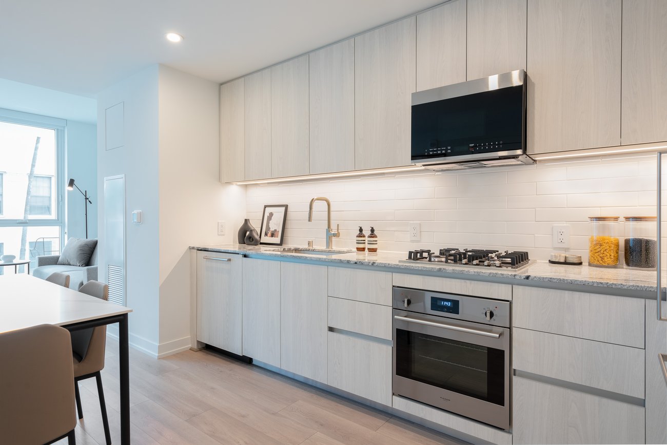 Modern kitchen with light wood cabinets, built-in oven, gas cooktop, stainless steel microwave, and sink. White tile backsplash, countertop with jars of pasta and utensils, and dining area visible on the left.