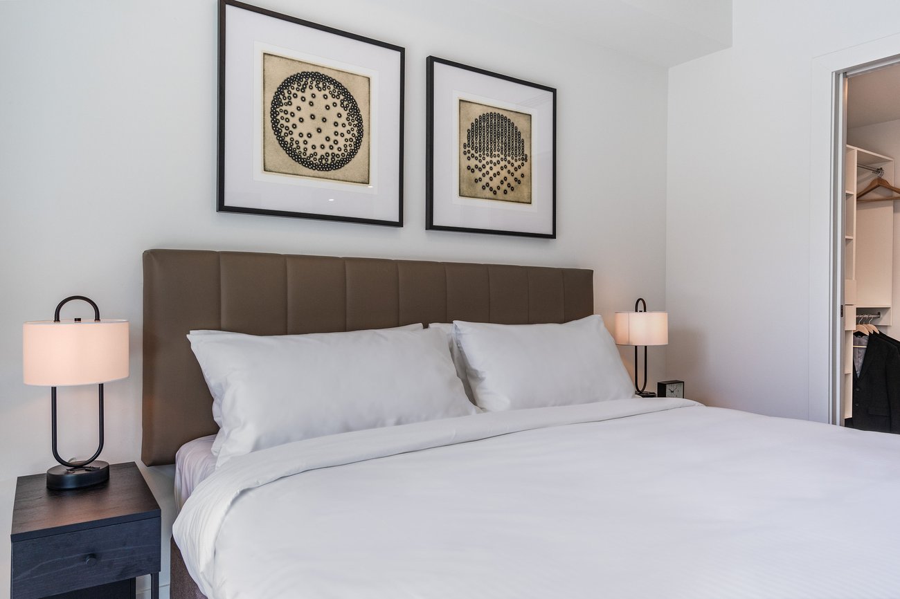 A neatly made bed with white bedding and a tan padded headboard sits between two black nightstands with matching lamps. Two framed abstract artworks hang above the headboard on a white wall.