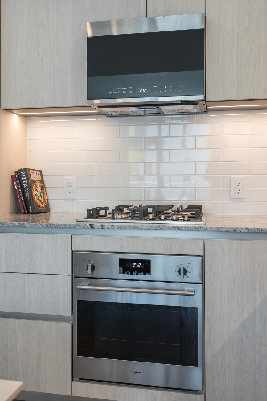 Modern kitchen with a built-in stainless steel oven and gas stovetop, light wood cabinets, white subway tile backsplash, and cookbooks on the countertop next to the stove.