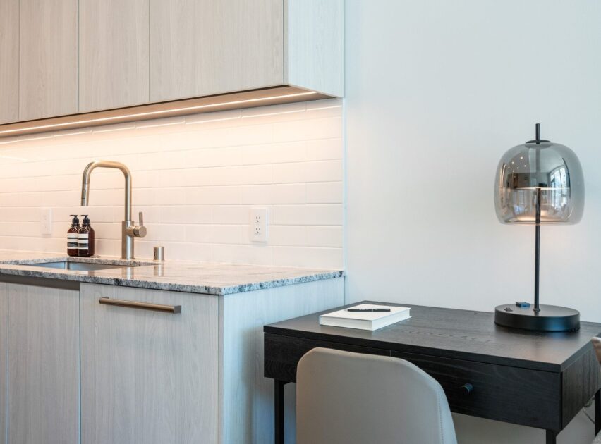 A modern kitchenette with a light wood finish, white tile backsplash, and a stainless steel faucet next to a small dark desk with a book and a glass table lamp.
