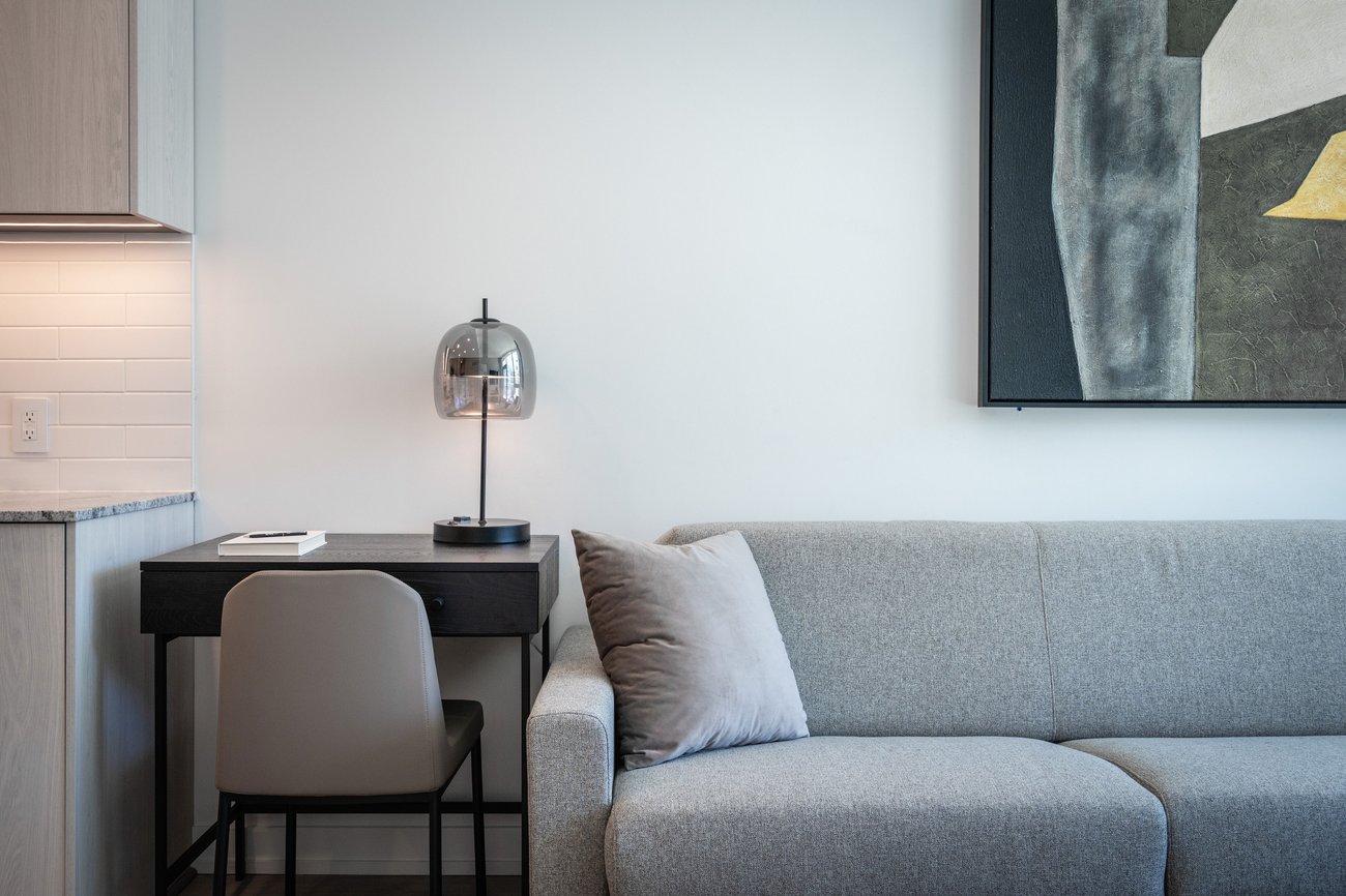A modern living space featuring a gray sofa with a light gray pillow, a small black desk with a gray chair, a glass lamp, an open notebook, and abstract wall art on a plain white wall.