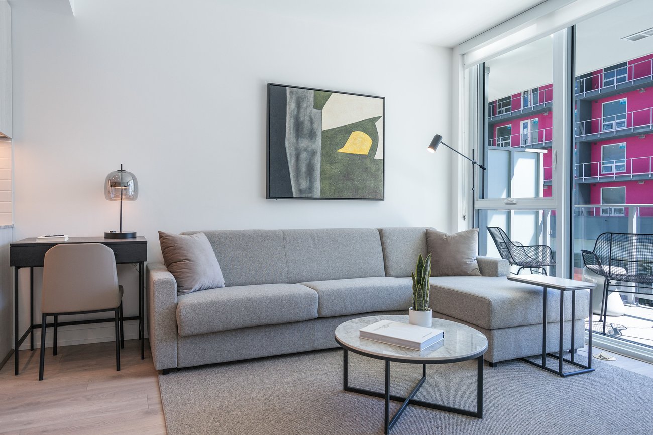 Modern living room with a gray sofa, round coffee table, small desk and chair, abstract wall art, floor lamp, and large windows with a view of a red building and balcony outside.