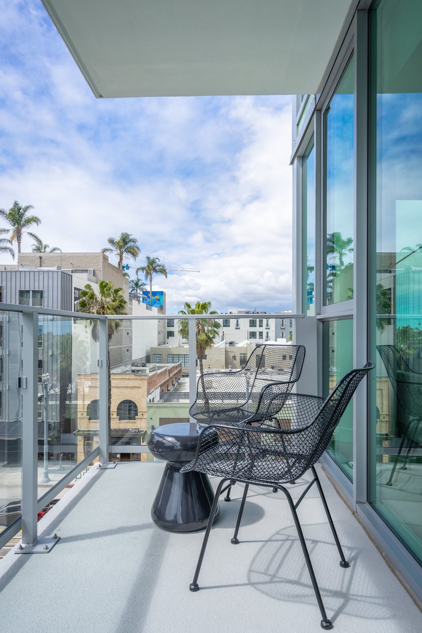A modern balcony with glass railings, two black wire chairs, and a small black table overlooks a cityscape with palm trees, buildings, and a partly cloudy sky.
