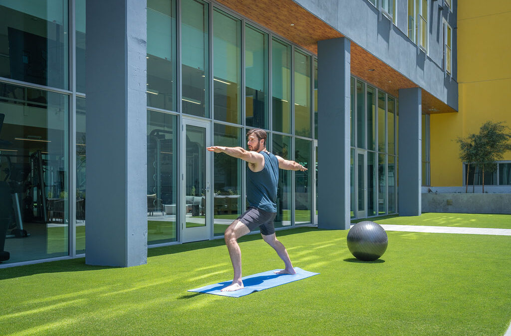 A man practices yoga outdoors on a mat, doing a warrior pose on green grass near a modern glass building. An exercise ball sits nearby.