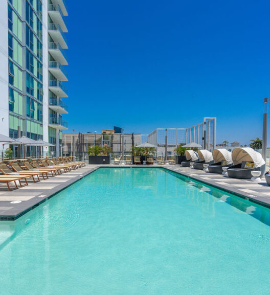 A modern rooftop pool with clear blue water, lined with wooden lounge chairs and shaded cabana beds, next to a tall glass building under a bright blue sky.