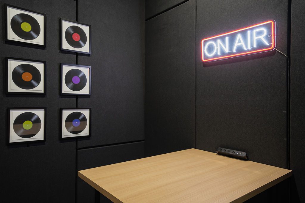 A small studio room with black soundproofed walls, a wooden table, a neon ON AIR sign, and six framed vinyl records arranged in two columns on the wall.