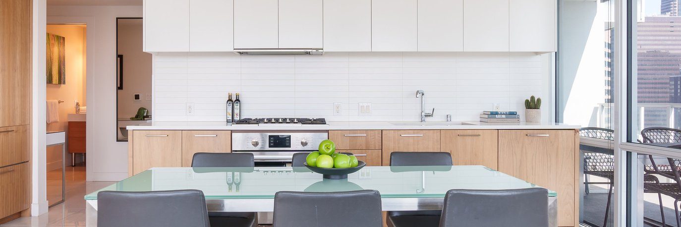 Modern kitchen with wood and white cabinets, a built-in oven, and a glass dining table with gray chairs. A black bowl of green apples sits on the table. Large windows reveal outdoor seating and city buildings.