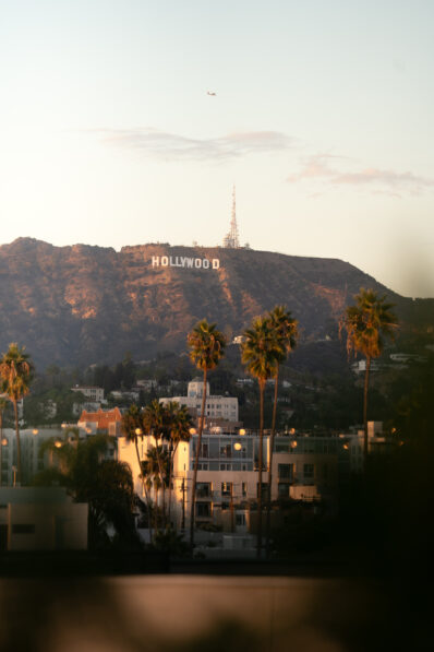 The Hollywood Sign sits atop a sunlit hill, with palm trees and buildings in the foreground under a clear sky; a small airplane is visible above the sign.