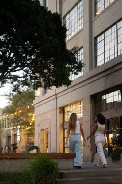Two women in light-colored outfits walk up steps toward a modern building with large windows, surrounded by greenery and string lights, during the evening.
