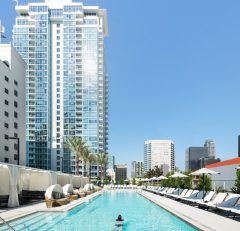 A person swims alone in a long outdoor pool lined with lounge chairs and cabanas, surrounded by tall modern skyscrapers under a clear blue sky.